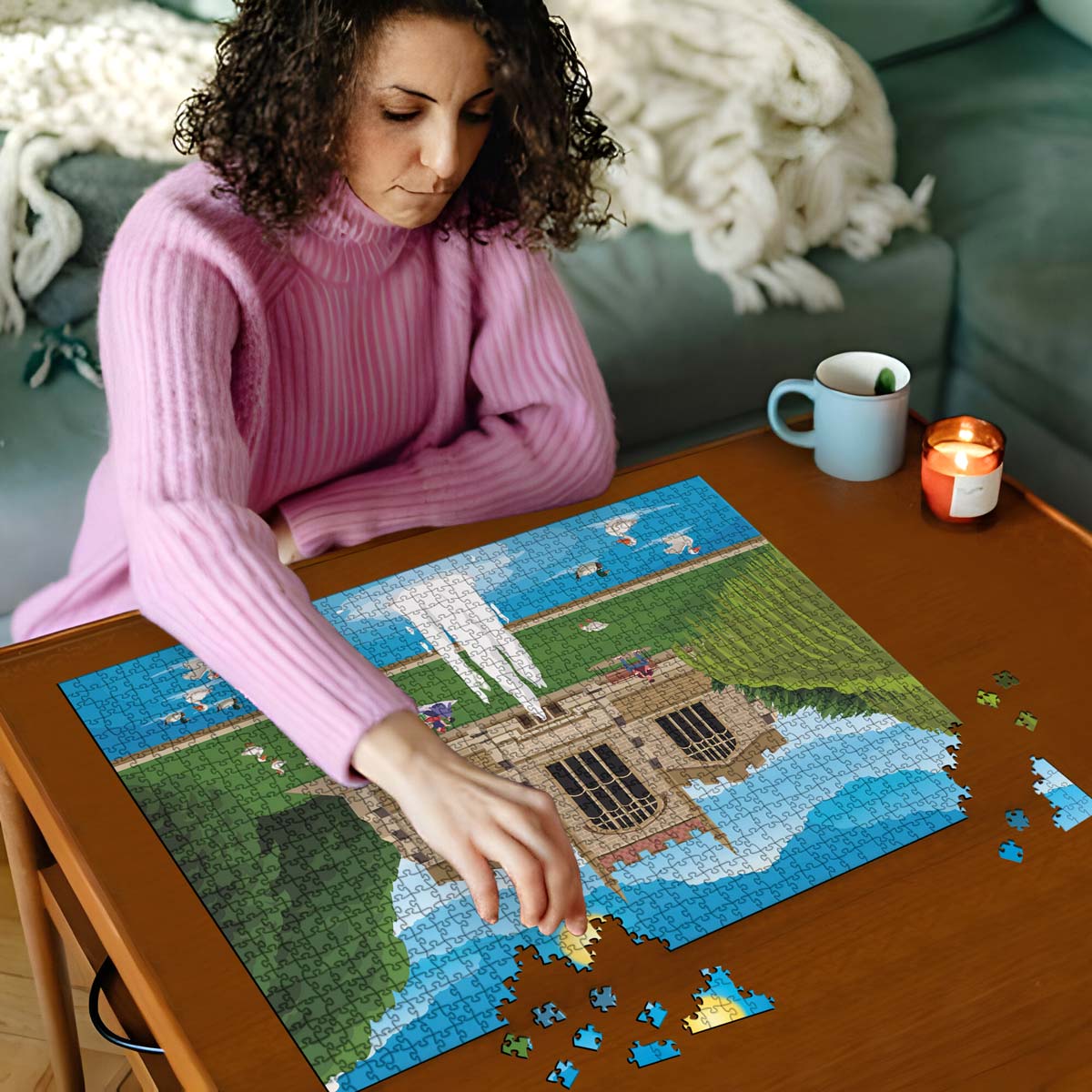 Woman in pink sweater working on a Hitchin St Mary's Church jigsaw puzzle on a wooden table with a cup and candles in the background