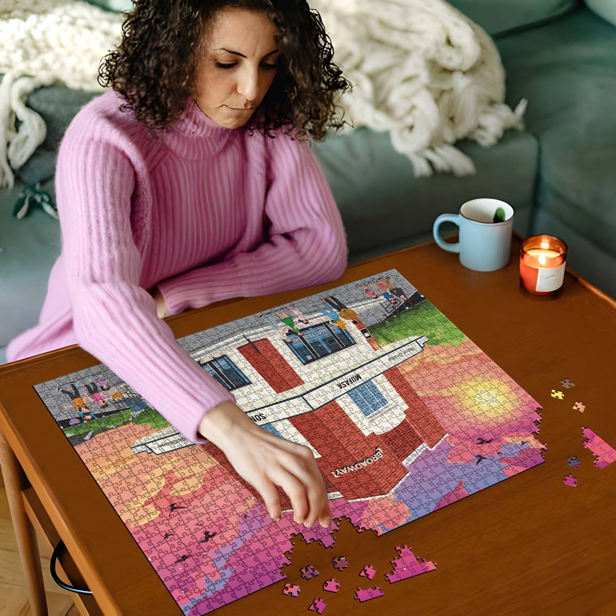 Woman in pink sweater working on a Letchworth Broadway Cinema jigsaw puzzle on a wooden table with a cup and candles in the background