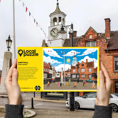 Hands holding "local Puzzle" box for Ampthill Town Centre jigsaw puzzle in front of the actual location 