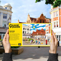 Person holding "local Puzzle" box for Bedford Town Centre jigsaw puzzle in front of the actual location 
