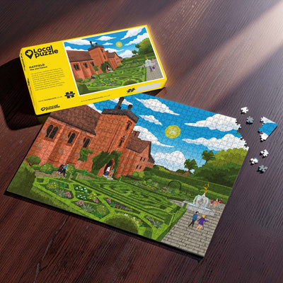 A completed Hatfield Old Palace jigsaw puzzle of the palace and its gardens, displayed on a dark wooden table next to its yellow packaging box and a few loose puzzle pieces.