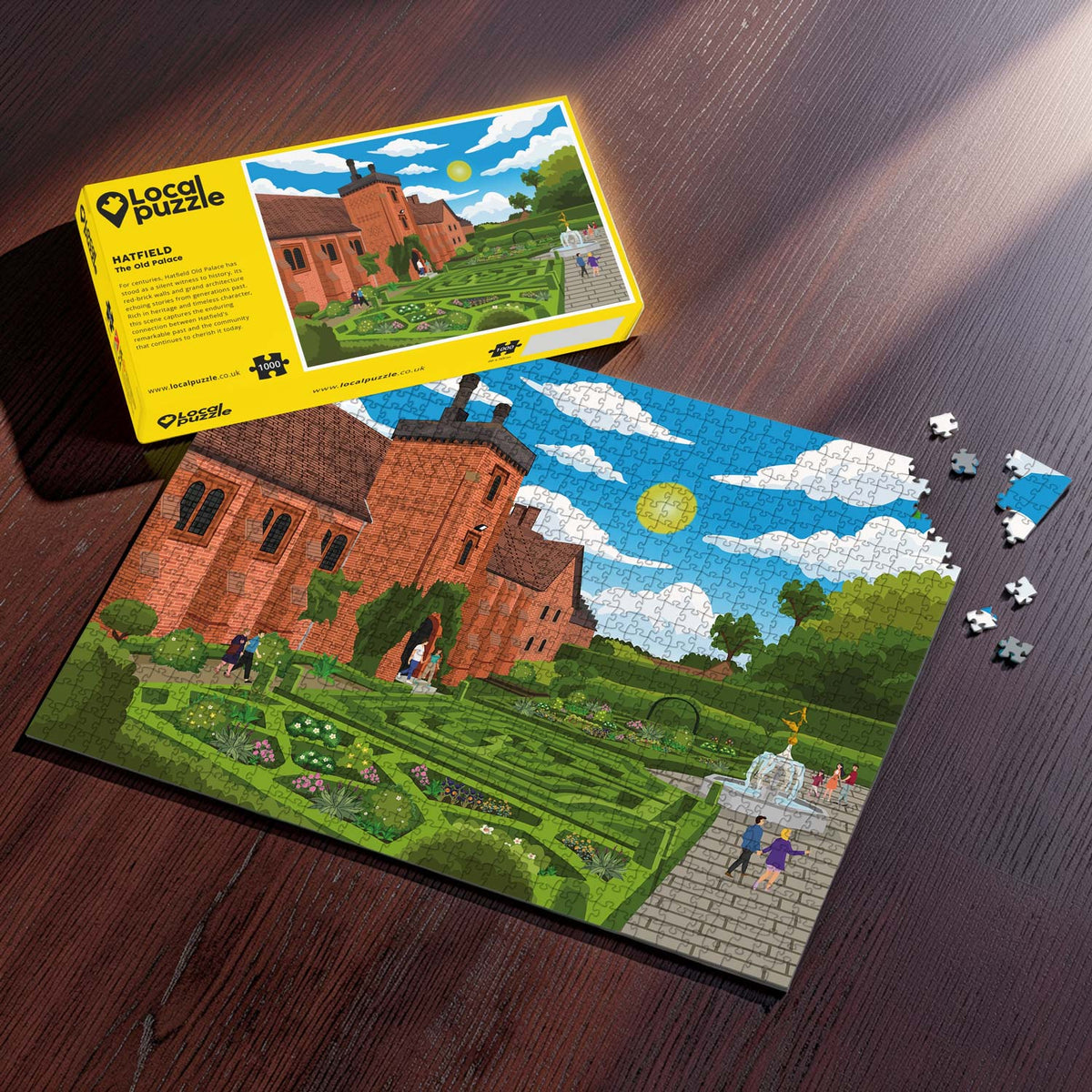 A completed Hatfield Old Palace jigsaw puzzle of the palace and its gardens, displayed on a dark wooden table next to its yellow packaging box and a few loose puzzle pieces.