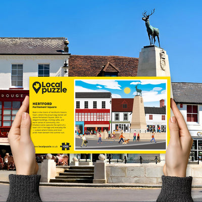 Person holding "local Puzzle" box for Hertford Parliament Square jigsaw puzzle in front of the actual location 