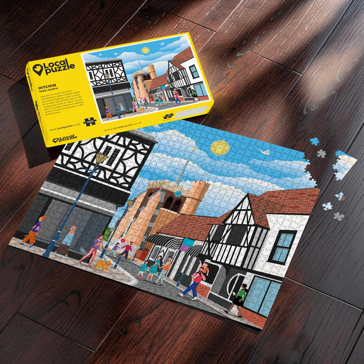 A completed Hitchin Town Centre jigsaw puzzle showing a colorful town scene with buildings, people, and a blue sky, placed on a dark wood floor next to its yellow Local Puzle packaging box.