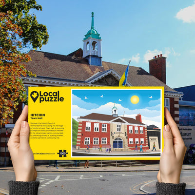 Person holding "local Puzzle" box for Hitchin Town Hall jigsaw puzzle in front of the actual location 