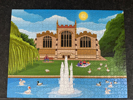 A completed jigsaw puzzle showing a colorful illustration of St Mary's Church in Hitchin with arched windows, green trees, a blue pond with ducks, and a fountain in the foreground, photographed against a dark textured background.