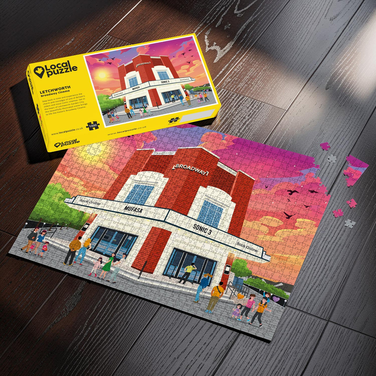 A completed Letchworth Broadway Cinema jigsaw puzzle showing a brick building art-deco cinema with people and a sunset sky, displayed on a dark wood surface next to its yellow Local Puzle box.