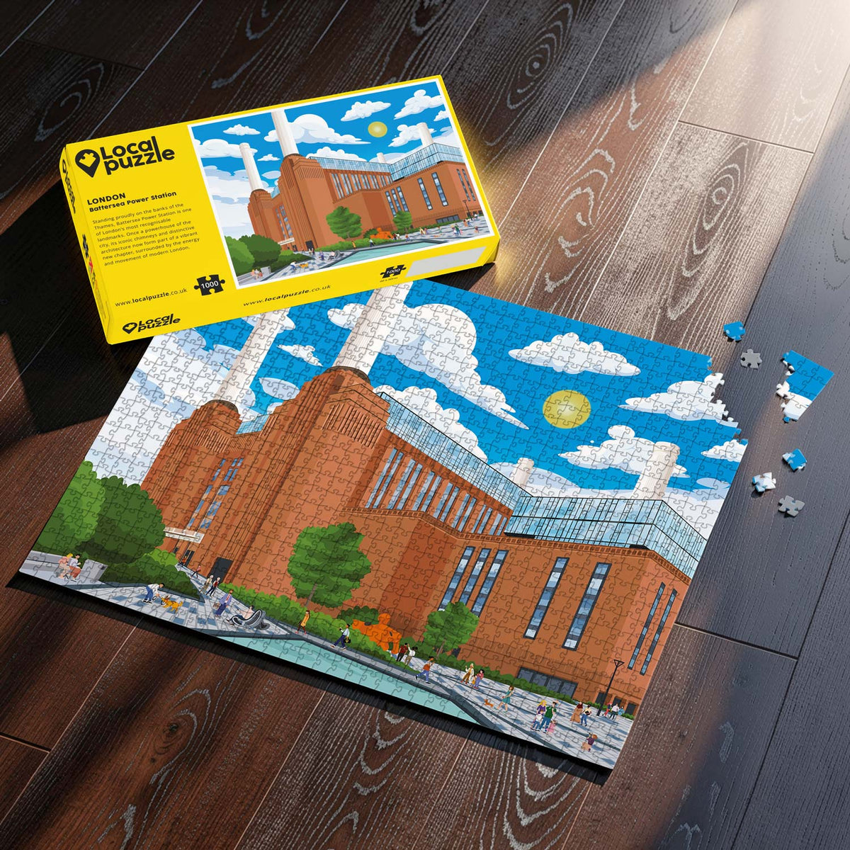 A completed London Battersea Power Station jigsaw puzzle and its yellow box are shown on a dark wood floor. The puzzle depicts a colorful scene with blue sky, white clouds, green trees, and small people walking in front.
