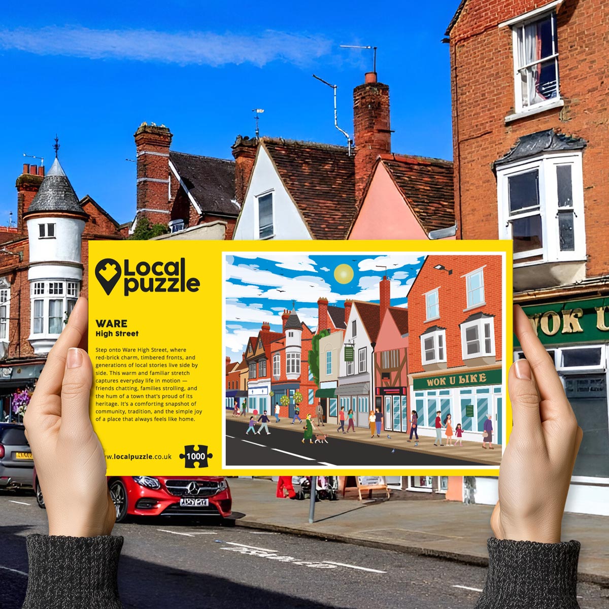 Person holding "local Puzzle" box for Ware High Street jigsaw puzzle in front of the actual location 