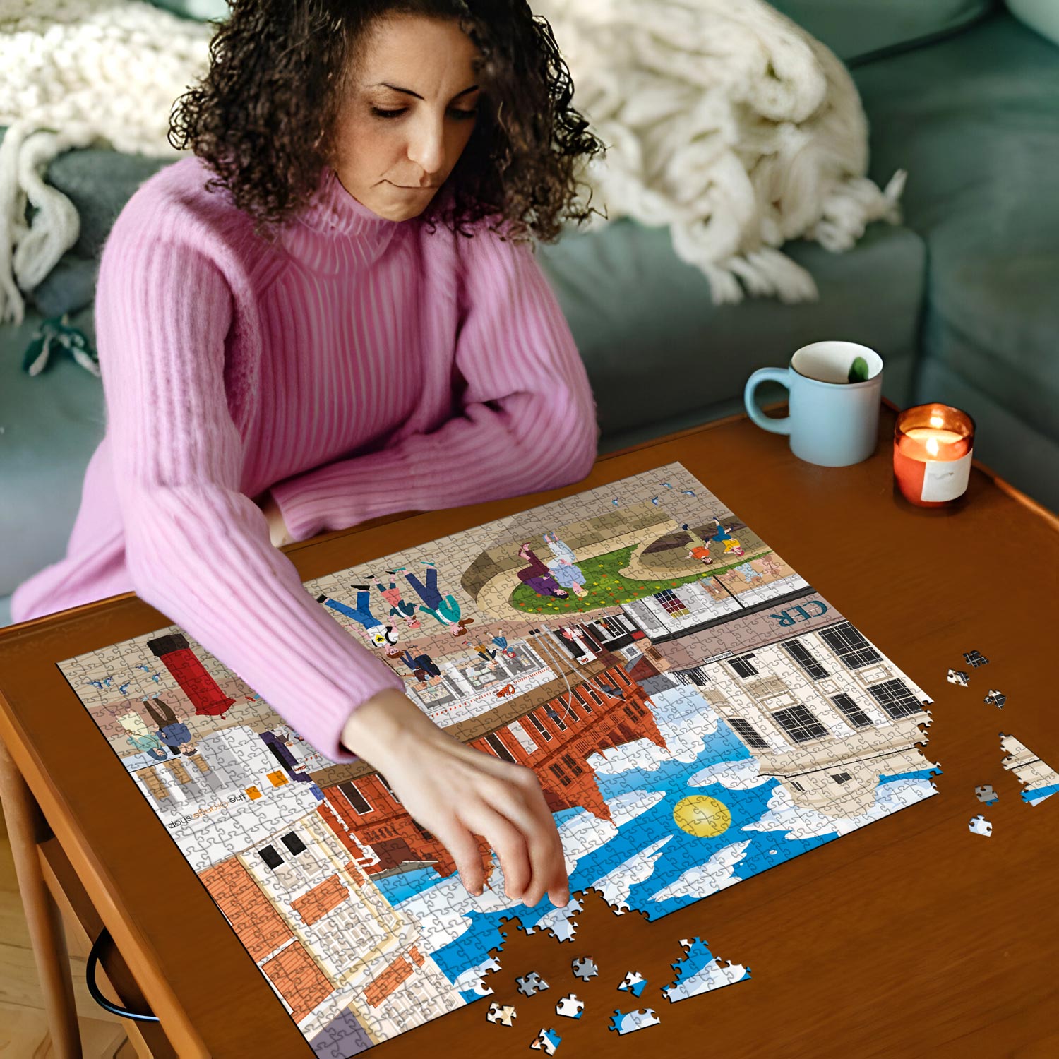 Woman in pink sweater working on a Bedford town centre jigsaw puzzle on a wooden table with a cup and candles in the background