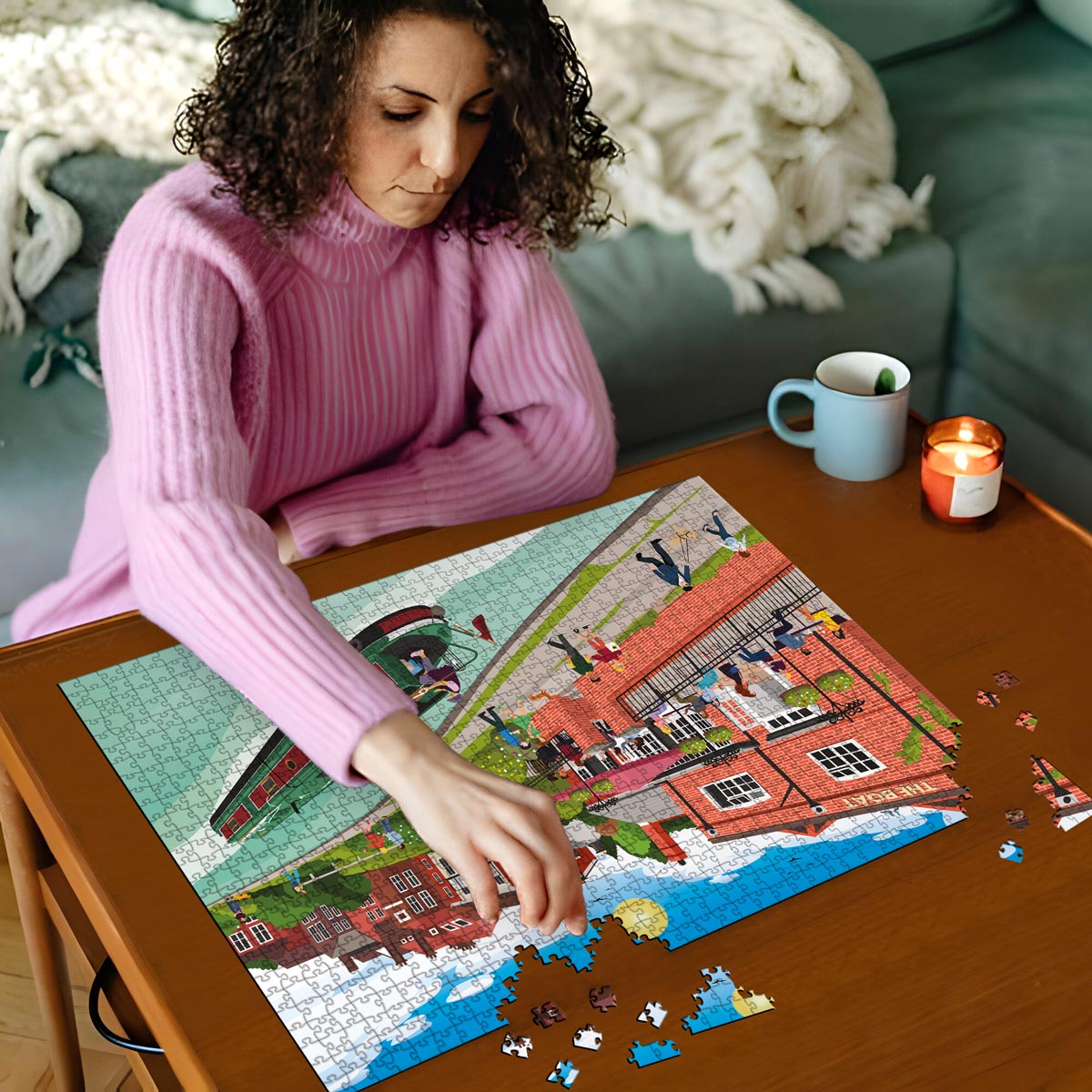 Woman in pink sweater working on Berkhamsted Grand Union Canal jigsaw puzzle on a wooden table with a cup and candles in the background