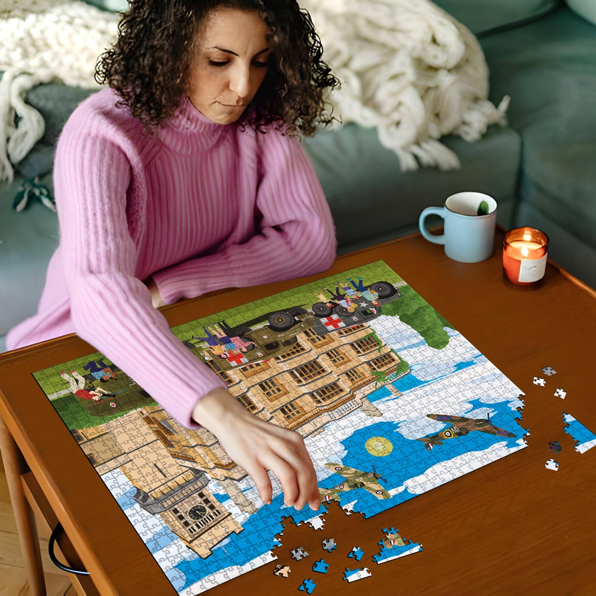 Woman in pink sweater working on a Biggleswade Shuttleworth House jigsaw puzzle on a wooden table with a cup and candles in the background