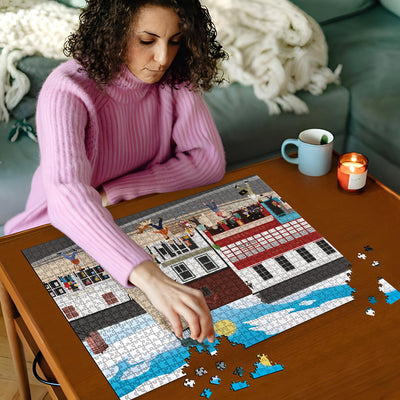 Woman in pink jumper working on our Hertford Parliament Square jigsaw puzzle on a wooden table with a cup and candles in the background