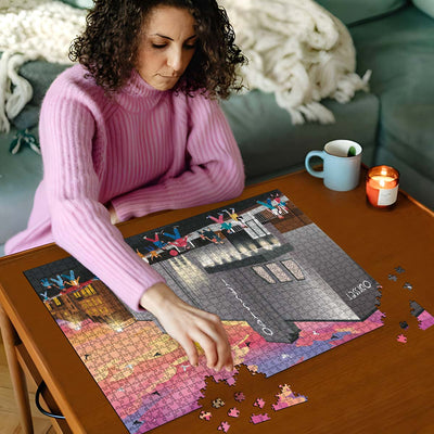 Woman in pink sweater working on a St Albans Odyssey Cinema jigsaw puzzle on a wooden table with a cup and candles in the background