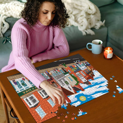 Woman in pink sweater working on a Ware High Street jigsaw puzzle on a wooden table with a cup and candles in the background