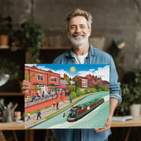 Man holding a completed Berkhamsted Grand Union Canal jigsaw puzzle of a canal scene with buildings and people.