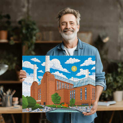 A smiling man with gray hair and a beard holds up a completed London battersea Power Station jigsaw puzzle showing a colorful illustration of the building under a bright blue sky with white clouds.