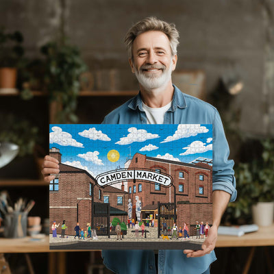 A smiling man with gray hair and a beard holds up a completed London Camdem Market jigsaw puzzle. The puzzle shows a blue sky with white clouds, a yellow sun, and a red-brown brick market hall with people walking in front of the main entrance. 
